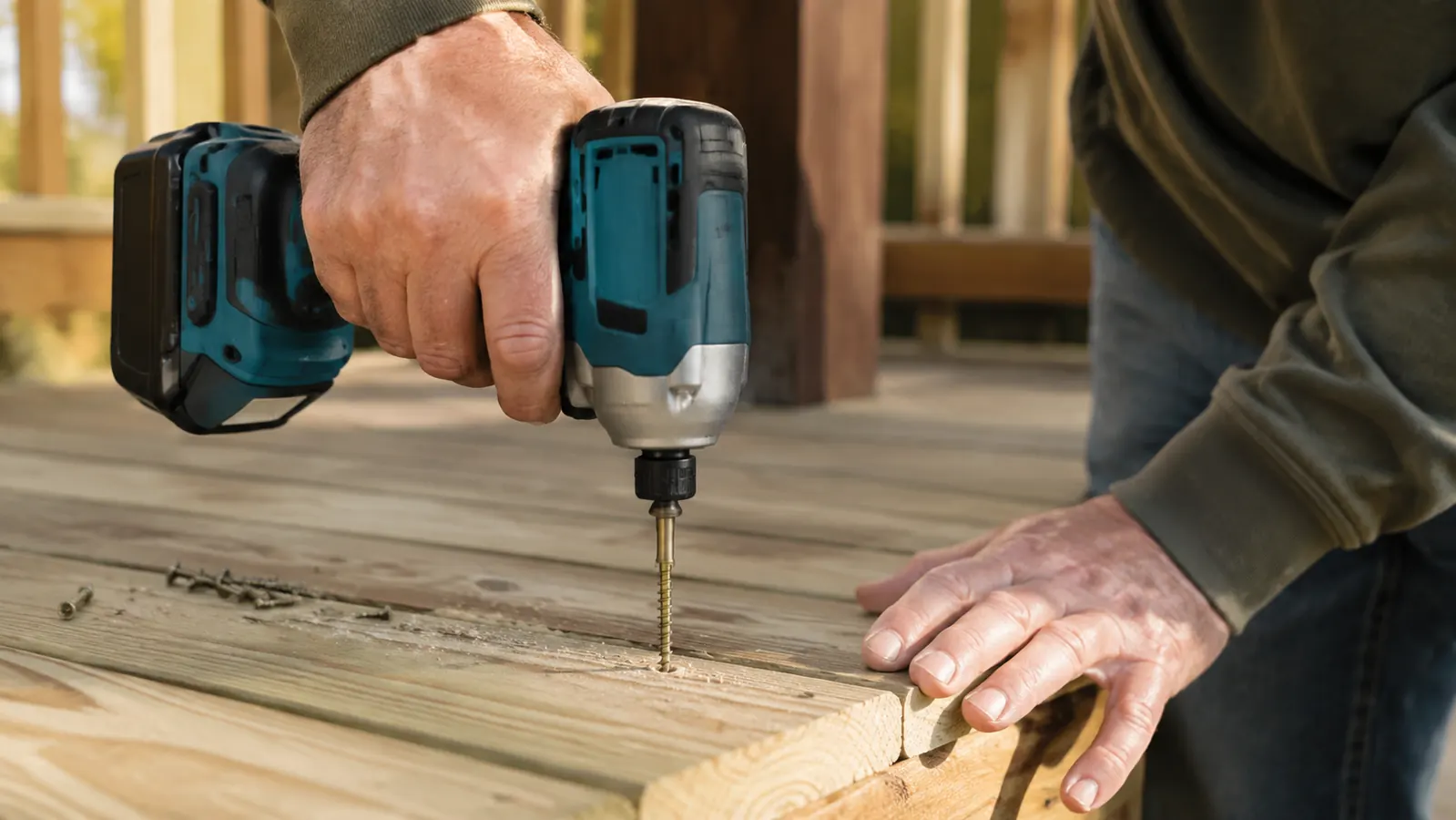 A hand driving a screw into a pressure-treated deck board with a cordless impact driver, sawdust and a few loose screws on the board, late afternoon light.