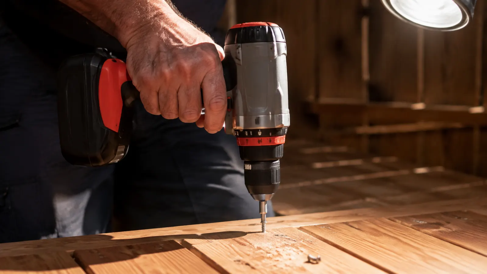 A hand gripping a cordless drill driving a screw into a cedar deck board, a bright work light overhead
