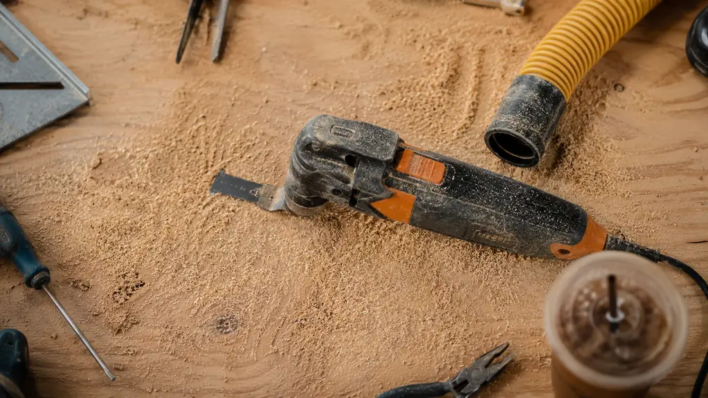 Close-up of a gloved hand gripping the oscillating tool on a workshop bench, dust collected around the base, a shop vac hose nearby