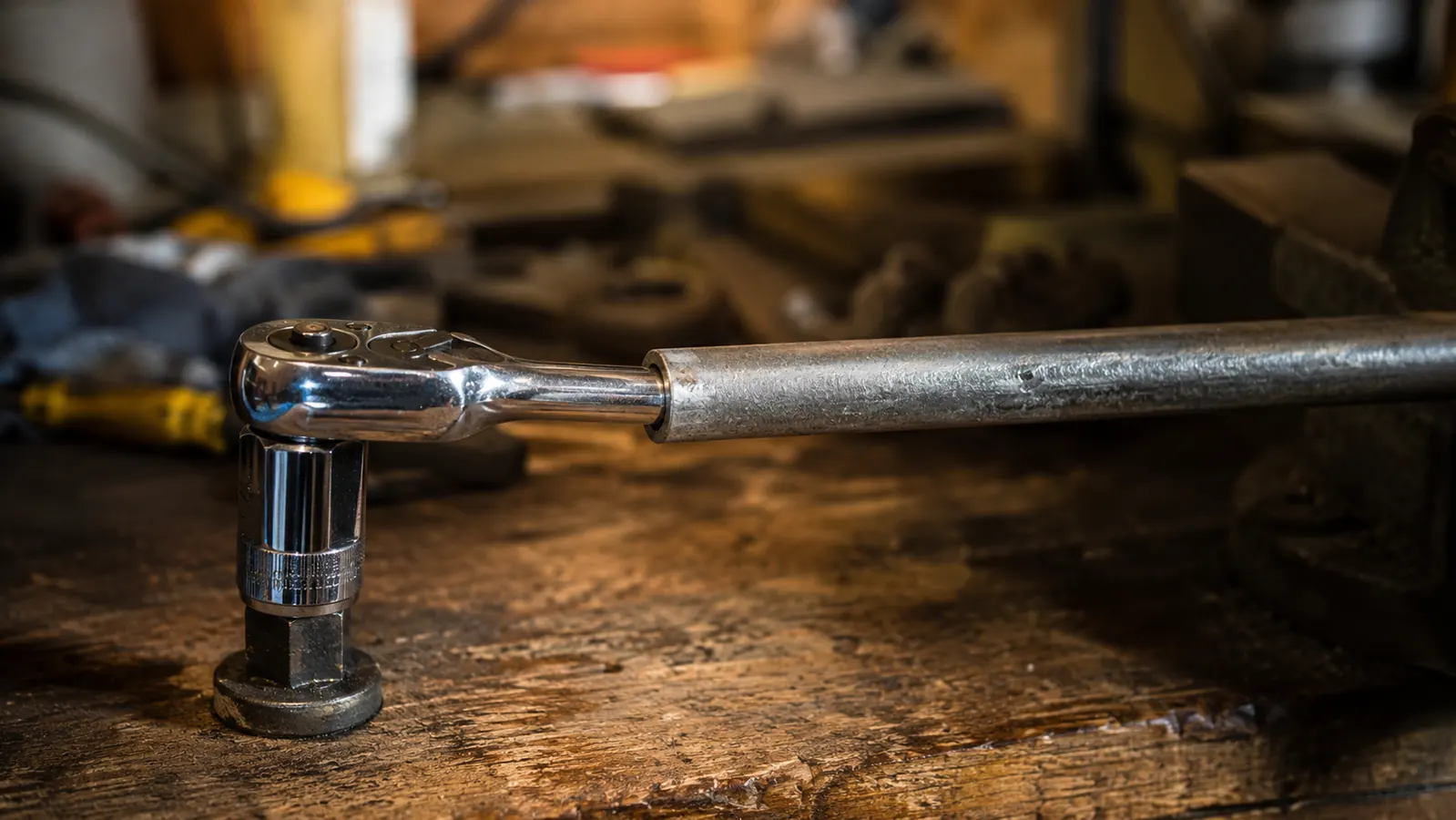 A chrome socket on a ratchet with a short steel cheater bar slipped over the handle, braced on a stubborn bolt on a workbench
