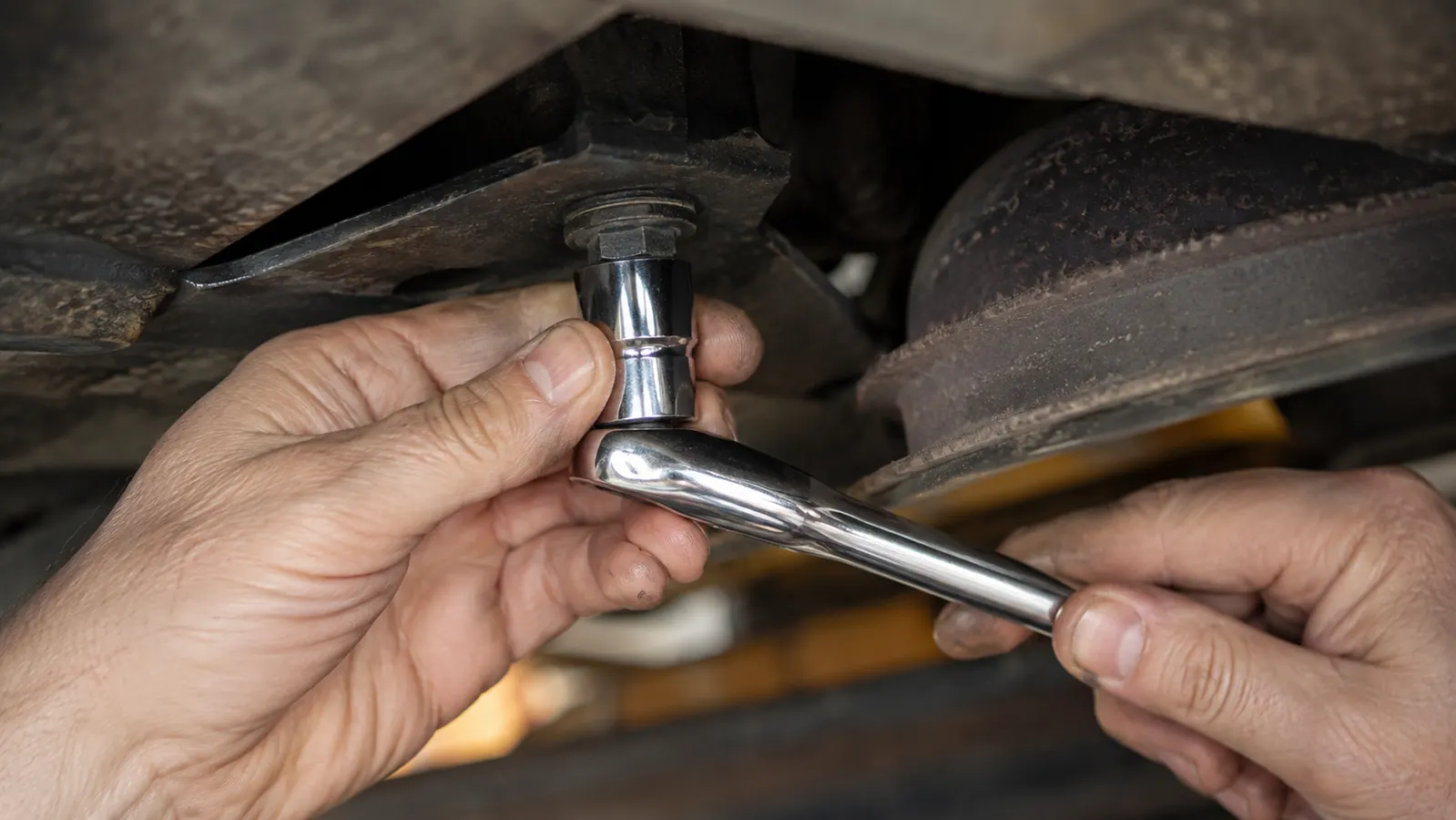 A hand fitting a chrome socket and pear head ratchet onto a bolt head under a workbench, close view with the tool angled into a tight space